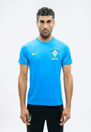 Bărbat stând cu fața înainte, purtând un tricou al echipei de fotbal Brazilia, de culoare albastră, și pantaloni negri cu emblema Braziliei, pe un fundal alb neutru.