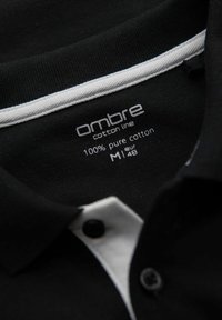 Svart polo-skjorta med vit krageaccent, tillverkad av 100% ren bomull; har varumärkesetikett och storleksinformation på insidan.