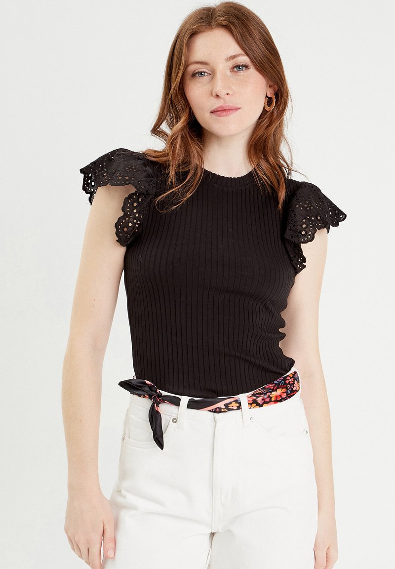 Top nero a costine con maniche in pizzo occhiellato a scallop, con un design aderente. Abbinato a jeans bianchi e una cintura in tessuto con motivo.