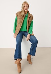 Beige gepolsterte Weste über einem grünen Strickpullover, kombiniert mit blauen Schlagjeans und beigen Ankle Boots. Das Model sitzt vor einem neutralen Hintergrund.