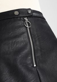 Czarna spódnica z ekoskóry z srebrnym zamkiem i dwoma okrągłymi metalowymi guzikami na pasku, prezentowana na manekinie.