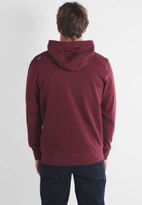 Sweat à capuche bordeaux en tissu texturé, avec une couture dans le dos, des poignets et un ourlet côtelés. Inclut un petit logo de la marque sur l'épaule.