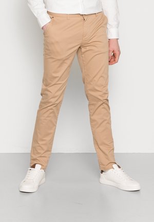 Uomo con pantaloni beige slim fit con le mani nelle tasche, abbinati a sneakers bianche e camicia bianca su sfondo semplice.