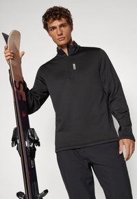 Sudadera de forro polar negra con cremallera y mangas largas, que presenta un cuello alto y un logo sutil, acompañada de esquís y fijaciones a un lado.