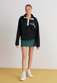 Pullover in pile nero con colletto bianco a metà zip, tasca frontale e polsini elasticizzati, abbinato a una gonna mini verde e sneakers bianche.