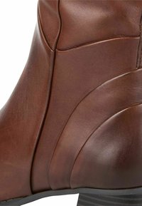 Bottine en cuir marron avec surface texturée, détails de couture courbés et talon empilé. Fini lisse avec un éclat subtil.