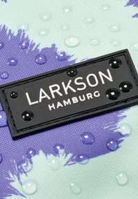 Közelkép egy fekete téglalap alakú címkéről, fehér "LARKSON HAMBURG" felirattal, kék és világos zöld szöveten vízcseppekkel.
