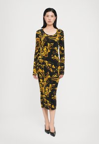 Svart långärmad klänning med gult blommönster, figursydd design, knälång, mjukt material, i kombination med svarta spetsiga klackskor.