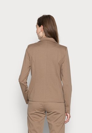 Kvinde med langt brunt hår står med ryggen mod og har en beige blazer og matchende bukser på foran en ensfarvet lys baggrund.