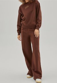 Sweat-shirt marron et pantalon large assorti en tissu doux, avec des poignets côtelés et un détail de logo discret sur le devant.