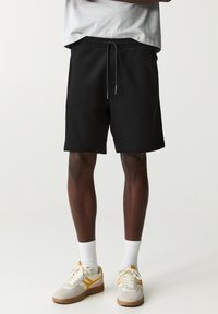 Schwarze Shorts mit elastischem Bund und Kordelzug, im lässigen Schnitt. Getragen mit weißen Socken und beigen Sneakers mit gelben Akzenten.