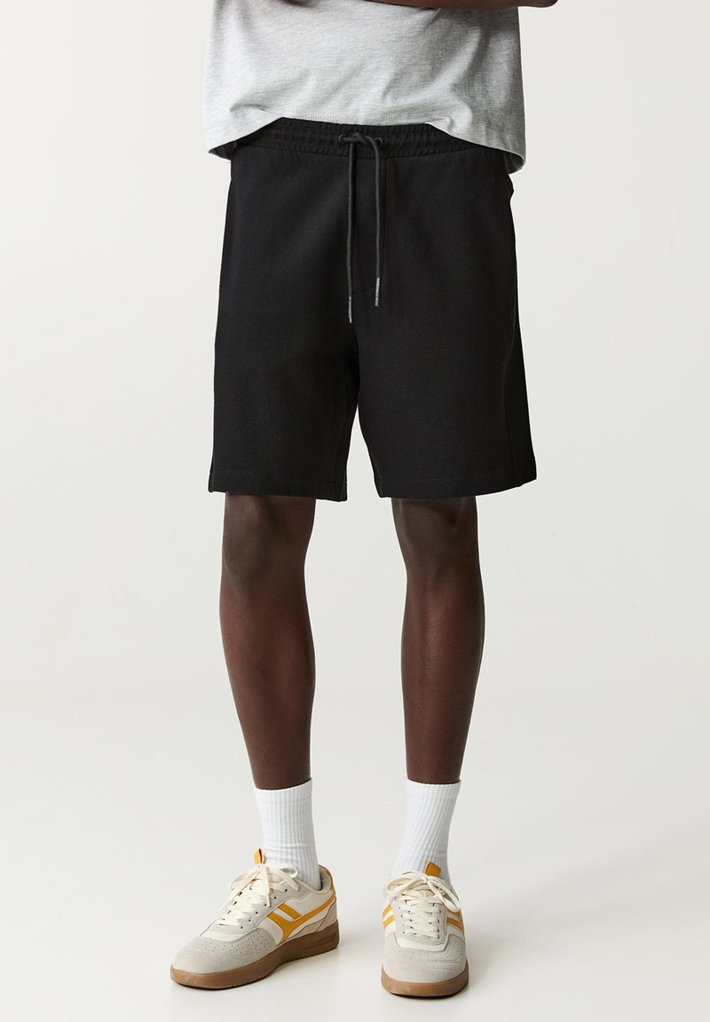 Schwarze Shorts mit elastischem Bund und Kordelzug, im lässigen Schnitt. Getragen mit weißen Socken und beigen Sneakers mit gelben Akzenten.