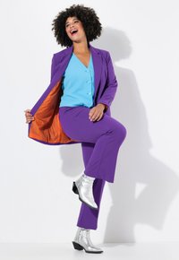 Abito viola con fodera arancione, top blu con bottoni e stivaletti argento alla caviglia. Presenta un blazer aderente e pantaloni a zampa.