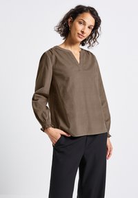 Donna con capelli ricci corti, indossa una blusa a maniche lunghe marrone e pantaloni neri, in piedi con una mano in tasca su uno sfondo bianco.