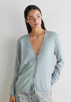 Frau mit langen dunklen Haaren, die eine hellblaue, geknöpfte Strickjacke und gemusterte graue Hose trägt und nach links an eine schlichte Wand schaut.