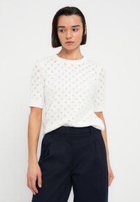 Λευκό πλεκτό πουλόβερ με κοντά μανίκια και διακοσμητικό μοτίβο διαμαντιού. Φοριέται πάνω από σκούρο μπλε παντελόνι. Ελαφρύ, υφασμάτινο ύφασμα με ριμπ γιακά.