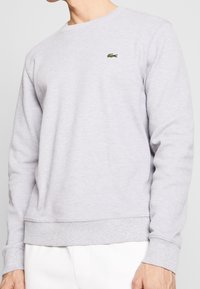 Uomo che indossa un felpa a maniche lunghe grigio chiaro con texture e un piccolo logo di coccodrillo verde sul petto, abbinato a pantaloni bianchi.