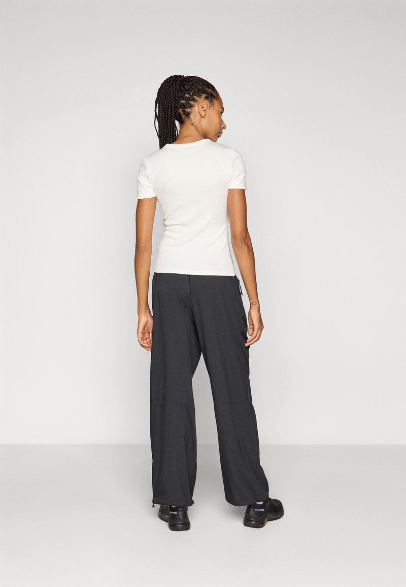 Weißes kurzärmeliges Shirt und lockere schwarze Hose, beide aus glattem Stoff. Die Hose hat eine Reißverschlusstasche und einen entspannten Schnitt. Schwarze Schuhe.