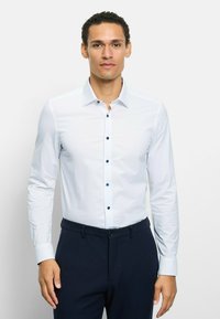 Weißes Langarmhemd mit klassischem Kragen und dunklen Knöpfen, kombiniert mit marineblauen Hosen; glatte Textur und tailliertes Design.