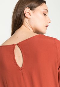 Blusa color ferrugem com um acabamento texturizado, apresentando um detalhe de buraco de fechadura na parte de trás com um pequeno fecho de botão.
