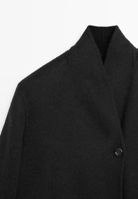 Czarny wełniany płaszcz o fakturowym wykończeniu, z kołnierzem z wyłogami i pojedynczym czarnym zapięciem na guzik. Minimalistyczny design z wyraźnie zarysowanym kształtem.
