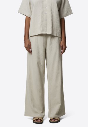 Kobieta ubrana w jasnobeżową, krótko rękawową koszulę o fakturze z guzikami oraz spodnie z szerokimi nogawkami, uzupełnione beżowymi sandałami na białym tle.