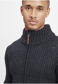 Mørkeblå strikket sweater med høj krave, der har strukturerede mønstre og sprøjt af rød, grøn og gul. Lynlås lukning med lædertræk.