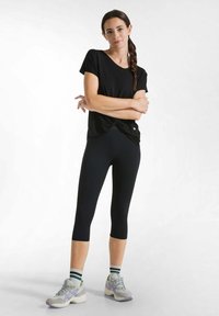 Maglietta nera a maniche corte con dettaglio a torsione, abbinata a leggings capri neri. Indossata con scarpe sportive grigie e calze a righe verdi.