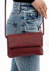 Burgundy leather crossbody bag with a fold-over flap, smooth texture, and subtle branding. Worn with high-waisted denim jeans.