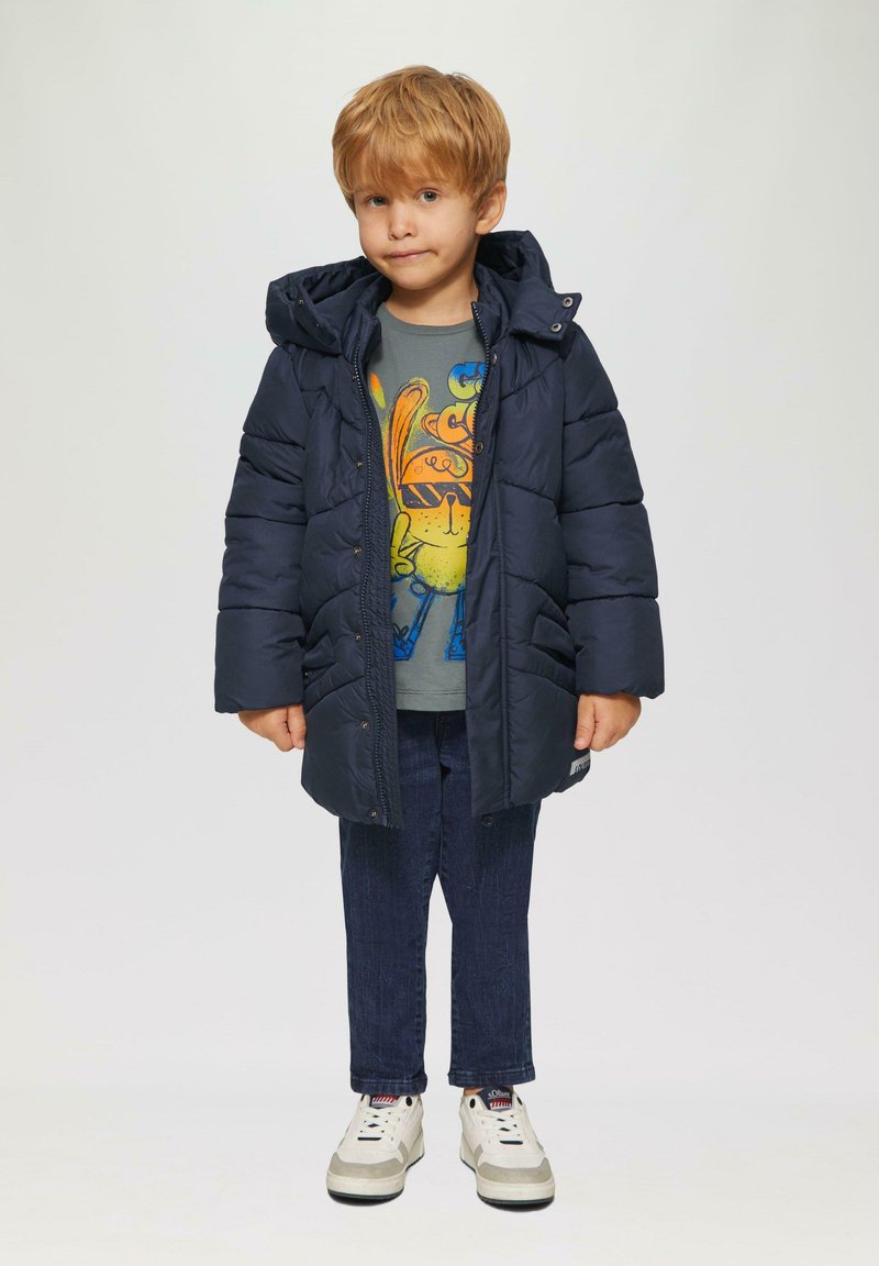 Marineblauer Pufferjacke mit Kapuze, gepolstertem Design. Getragen über einem grauen Shirt mit bunten Cartoon-Grafiken und einer dunklen Jeans. Weiße Sneakers.
