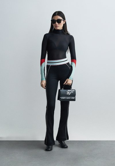 Γυναίκα που φοράει μαύρο ζιβάγκο με κόκκινες και μπλε πιέτες στα μανίκια, μαύρο παντελόνι με λευκές ραφές, μαύρες μπότες, γυαλιά ηλίου, κρατώντας μαύρη τσάντα με λευκό κείμενο.