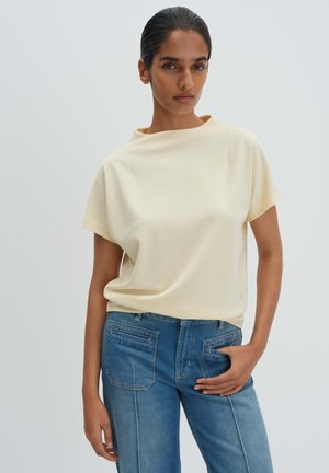 Giovane donna con capelli tirati indietro che indossa una blusa beige chiaro a maniche corte e jeans blu, in piedi contro uno sfondo semplice con una mano in tasca.