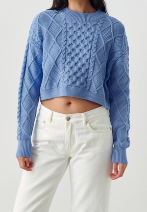 Modèle portant un pull court bleu clair en maille torsadée à manches longues et un jean blanc, sur un fond uni.