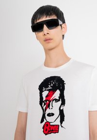 Gafas de sol negras de gran tamaño, camiseta blanca con un gráfico de una cara con un rayo rojo y "BOWIE" en texto rojo.