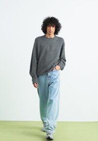 Maglione grigio oversize con maniche lunghe abbinato a jeans baggy azzurri. L'outfit include sneaker grigie e un tessuto strutturato.