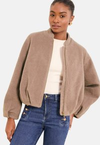 Chaqueta bomber beige de tela suave con cuello alto y cierre de cremallera. Presenta mangas abullonadas y un ajuste relajado. Combinada con vaqueros azules de talle alto.