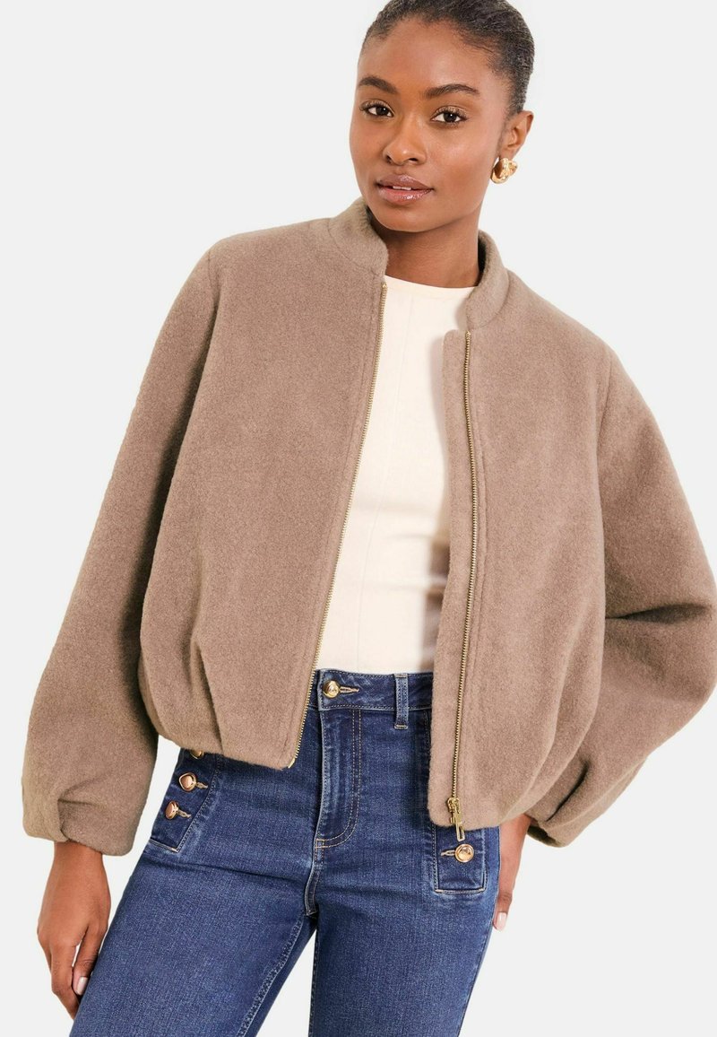 Chaqueta bomber beige de tela suave con cuello alto y cierre de cremallera. Presenta mangas abullonadas y un ajuste relajado. Combinada con vaqueros azules de talle alto.