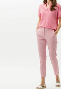 Kvinna iklädd en rosa kortärmad polo, rosa och vita randiga byxor med händerna i fickorna, samt vita espadrillos.