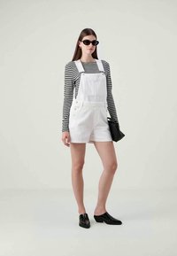 Kvinde iført en sort og hvid stribet langærmet skjorte, hvide korte overalls, sorte solbriller, sorte sko og holder en sort taske.