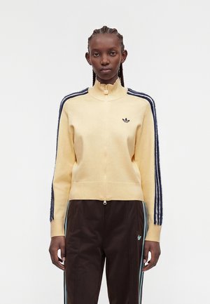 Joven mujer que lleva un suéter amarillo claro con cremallera y rayas azul oscuro en las mangas, y pantalones oscuros con rayas laterales azul claro.