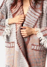 Textúrované vonkajšie oblečenie v šedej a broskyňovej farbe, s fringingovými okrajmi, jemnými vzormi a dvoma prednými vreckami so pruhovanými detailmi.