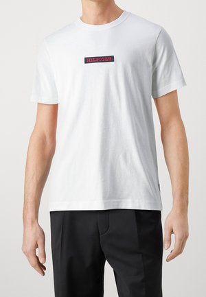 Mężczyzna nosi białą koszulkę na krótki rękaw z czarną, prostokątną naszywką z napisem "HILFIGER" w czerwonych literach, do tego czarne spodnie.