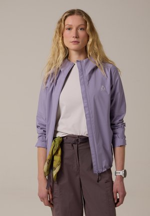 Joven con cabello rubio largo que lleva una chaqueta lila clara, camisa blanca, pantalones marrones, bufanda verde y reloj de pulsera blanco sobre un fondo neutro.