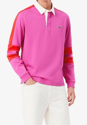Mann trägt pinkfarbenes Langarm-Poloshirt mit roten Streifen an den Armen und weißem Kragen, kombiniert mit weißen Hosen, Hand in der Tasche.
