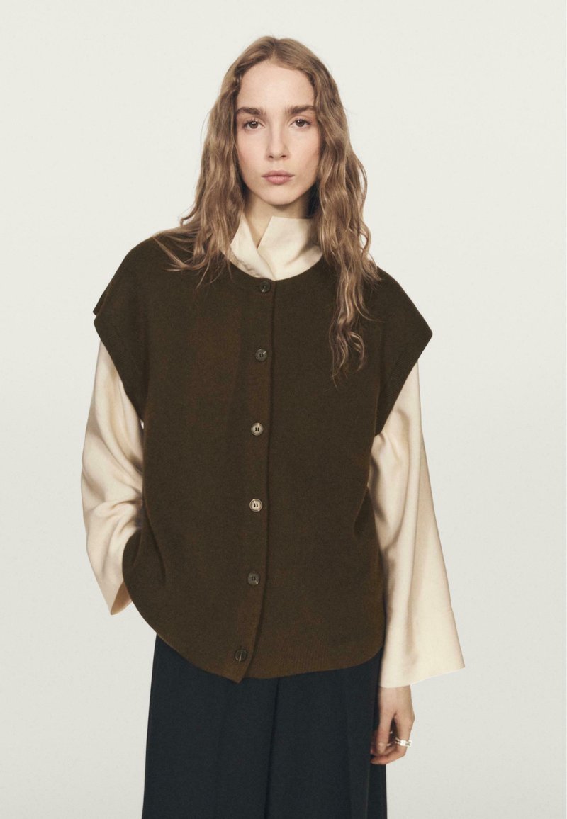 Dark green sleeveless sweater with a button-down front, layered over a light-colored, high-neck shirt, paired with dark pants.