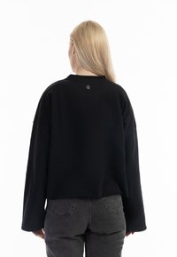 Bluza neagră scurtă cu mâneci lungi și guler ribbed. Are un mic detaliu cu logo în apropierea gulerului din spate. Este asortată cu blugi gri închis.