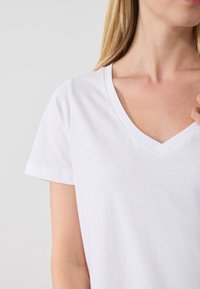 Maglietta bianca in cotone con scollatura a V e maniche corte, caratterizzata da una texture liscia e un design minimalista. Nessun motivo o logo visibile.