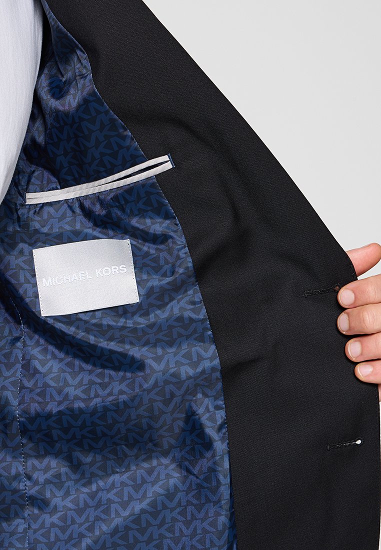 Hand die een open zwart colbert vasthoudt, met een donkerblauwe gestreepte voering, een binnenzak en een witte "MICHAEL KORS" label.