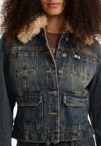 Giacca di denim con una finitura blu sbiadita, colletto in pelliccia sintetica, quattro tasche frontali e accenti in metallo argentato.