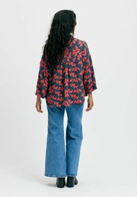 Välja skjorta med ett röd- och grått geometriskt mönster, ihopparad med utsvängda blå jeans. Mörkt hår faller över skjortans rygg.
